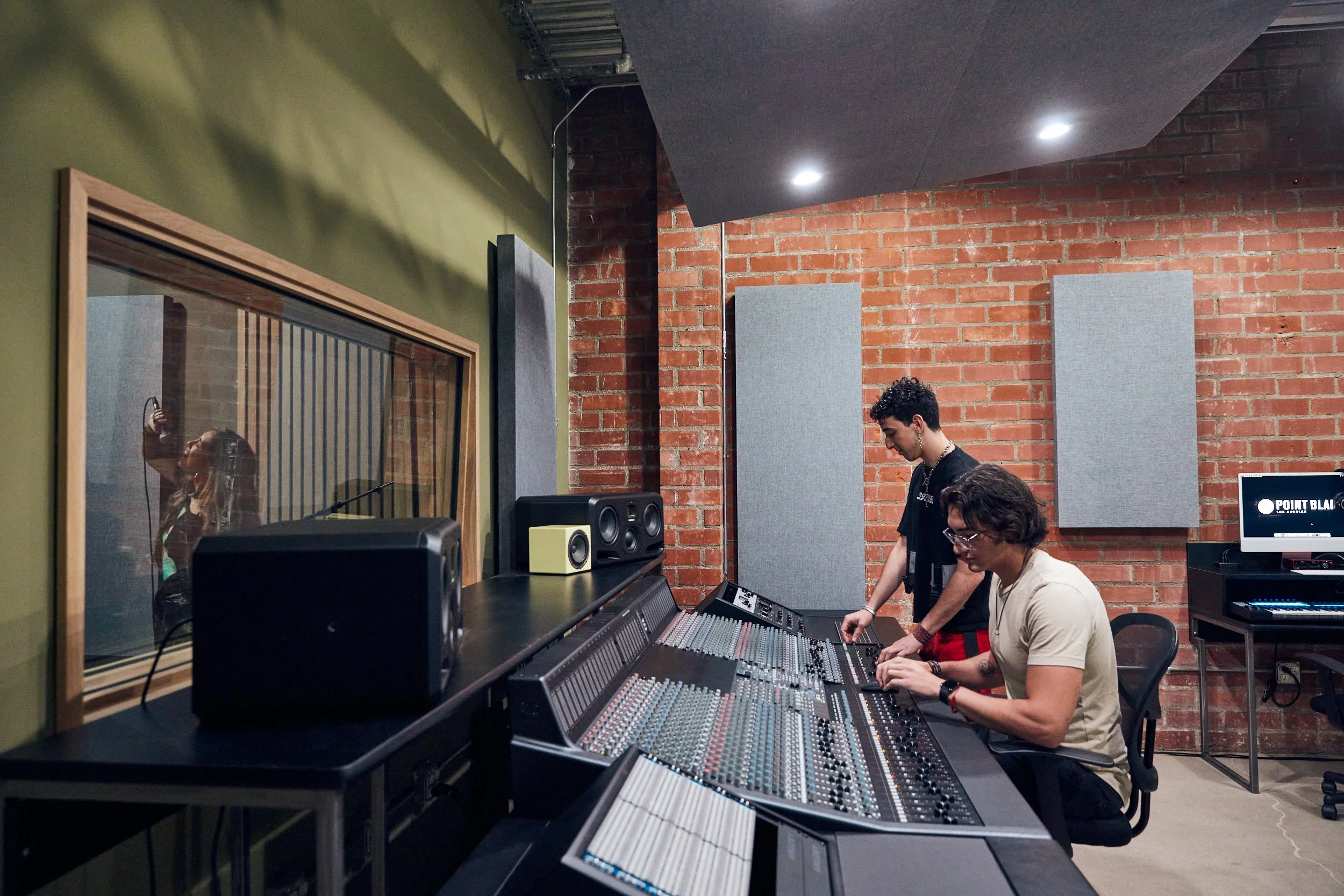 Two men at a large mixing console in a studio with brick walls and a vocal booth window.