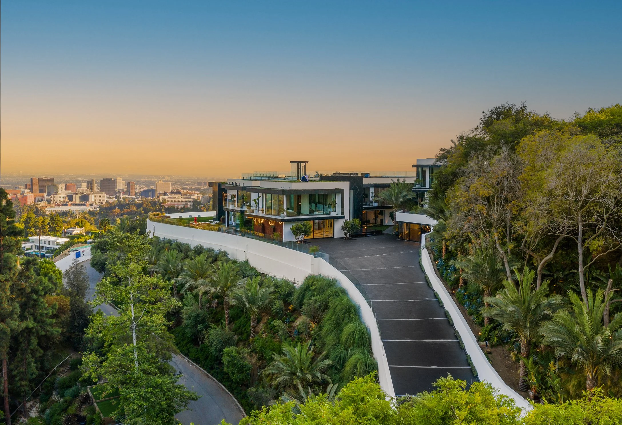 Multi-level glass hillside mansion above a curved driveway with a city skyline at golden hour.