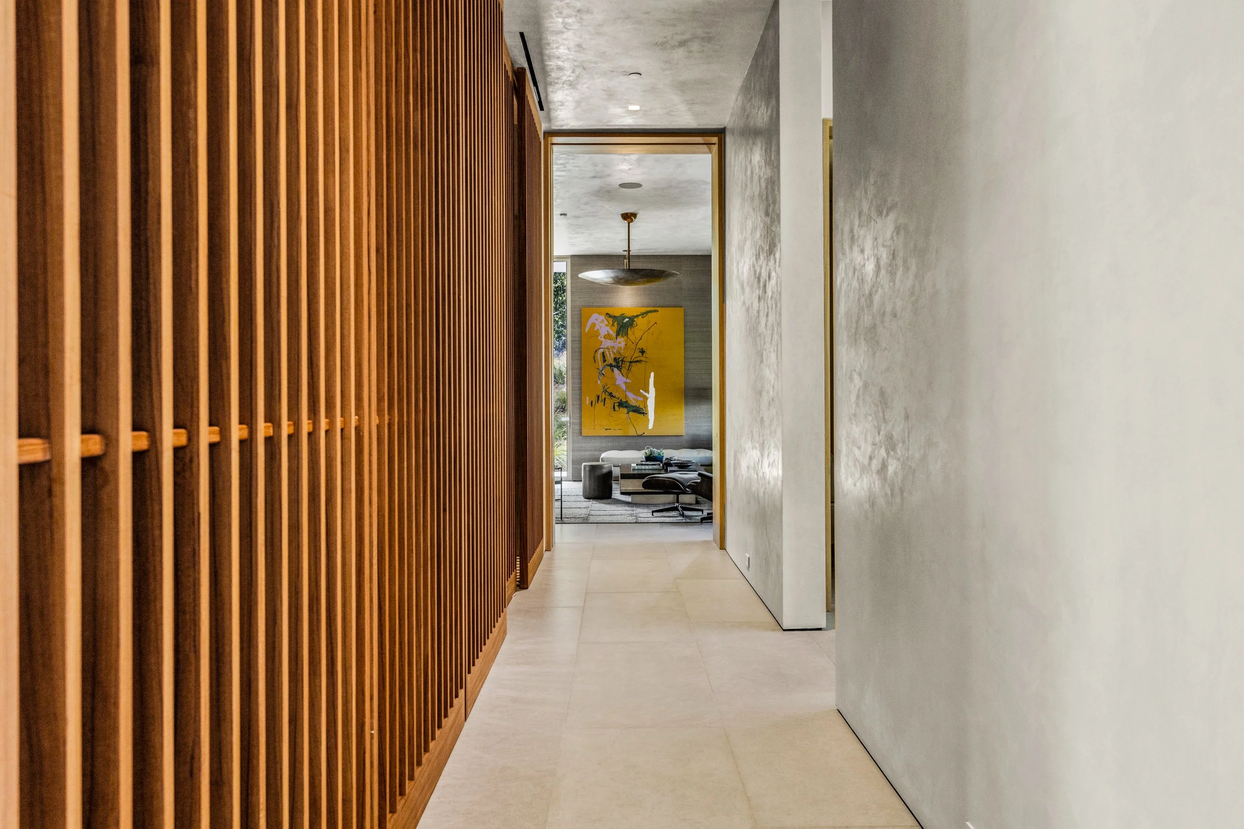 Hallway of vertical wood slats and plaster walls opening to a bright living room with yellow art.
