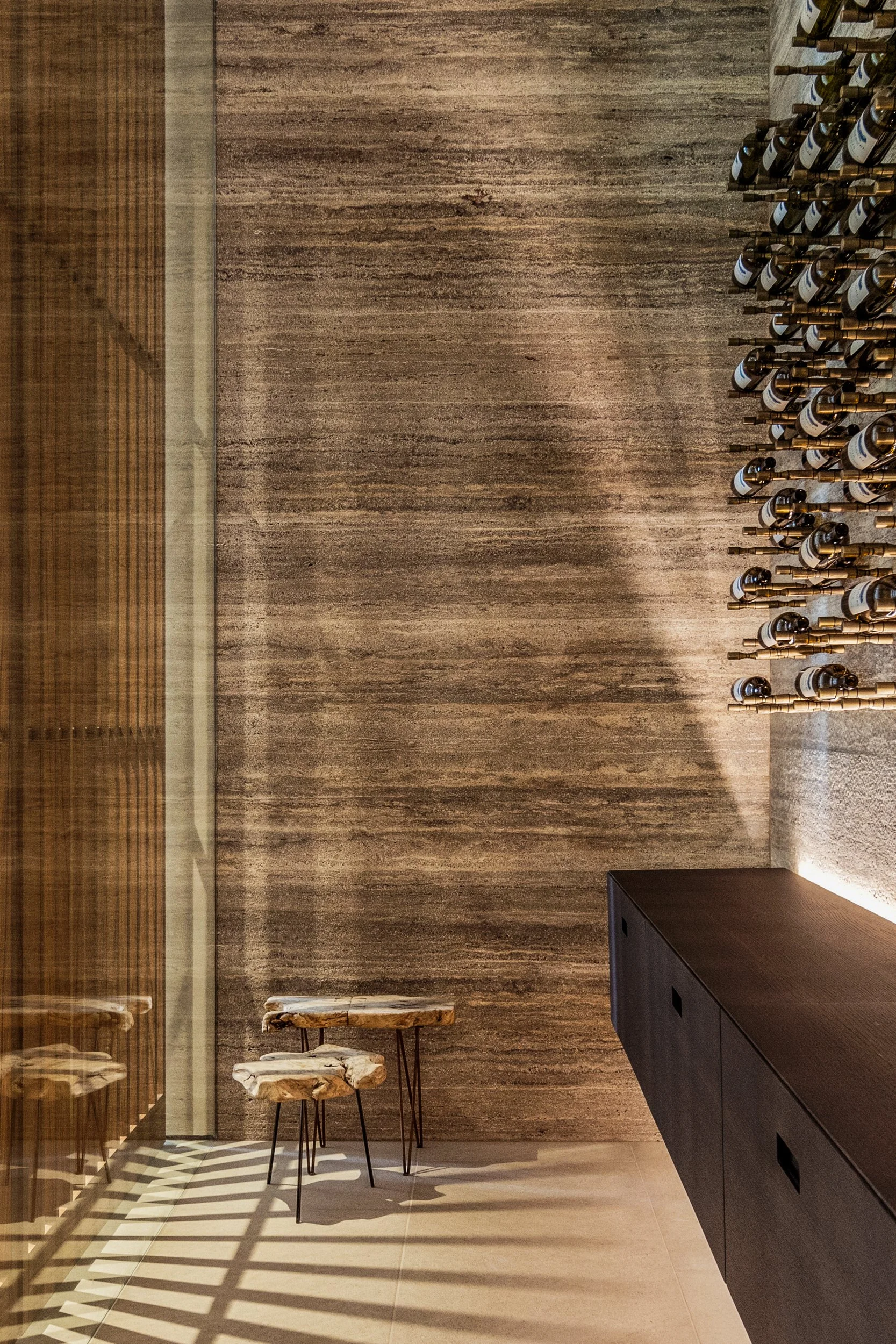 Wine room with a textured stone wall, floating dark cabinet, and peg-mounted bottles in soft light.