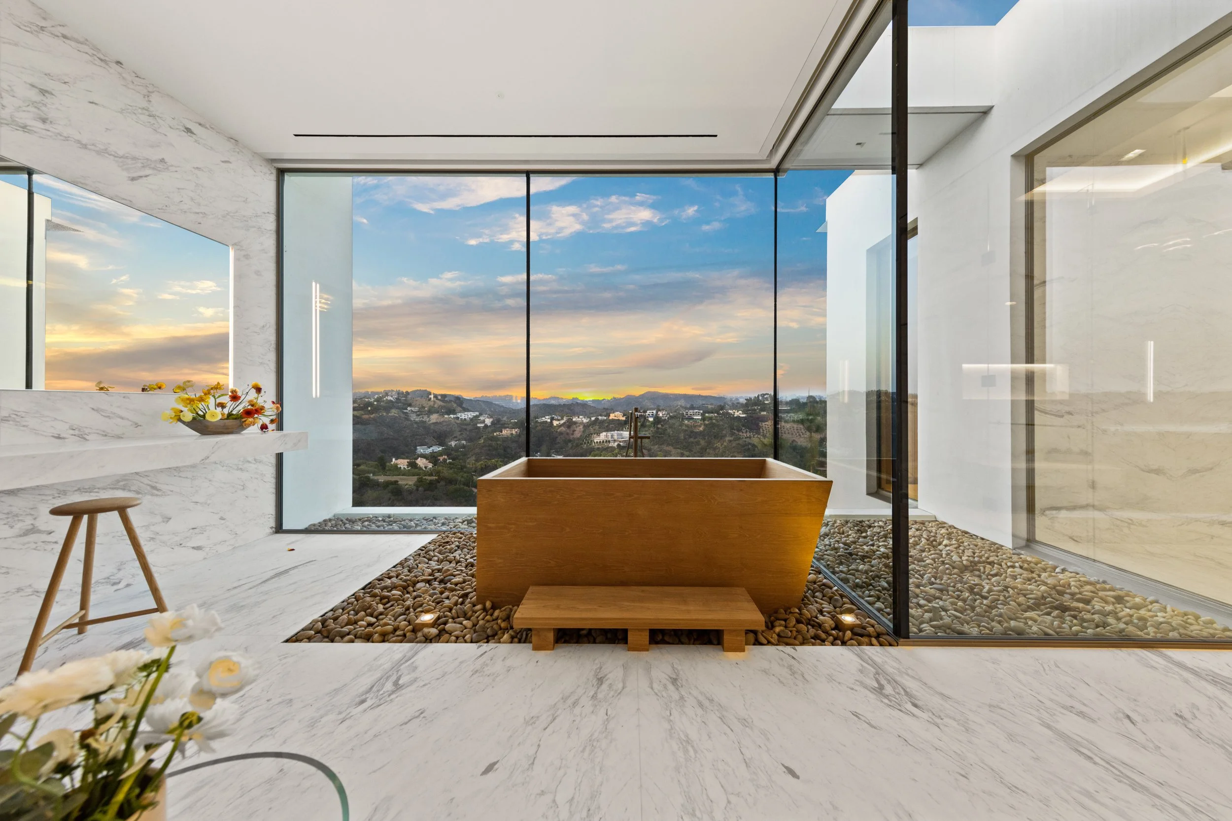 Marble bathroom with a wooden soaking tub on river rocks before sunset hillside windows.