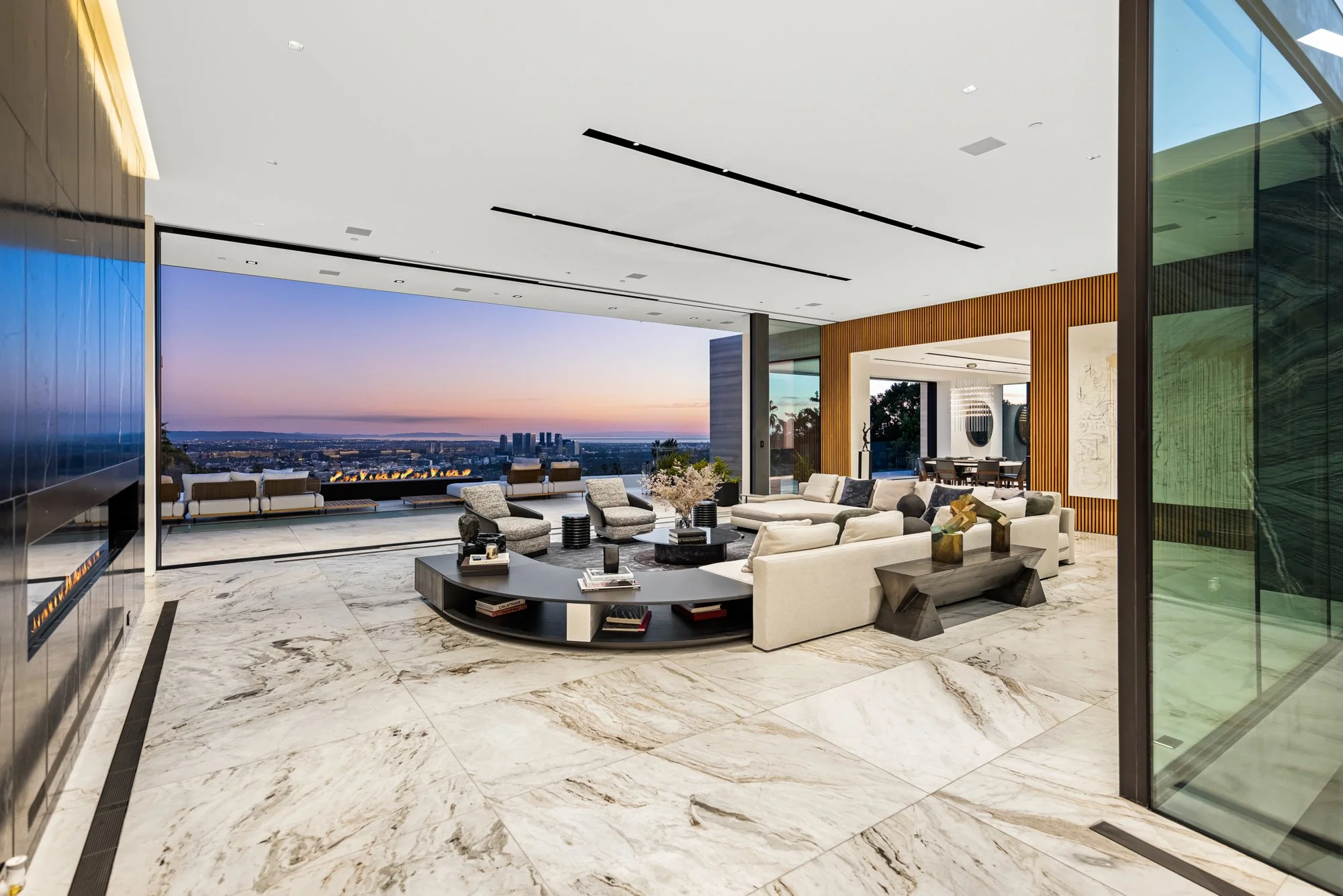 Open living room with a curved sofa and marble floors opening to a city terrace at dusk.
