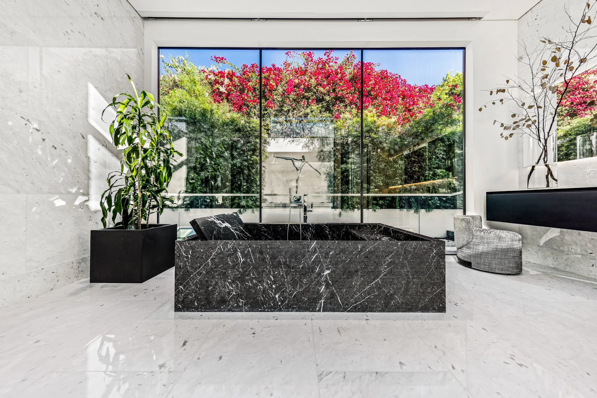 Black marble freestanding tub on white marble floors before a window of pink bougainvillea.
