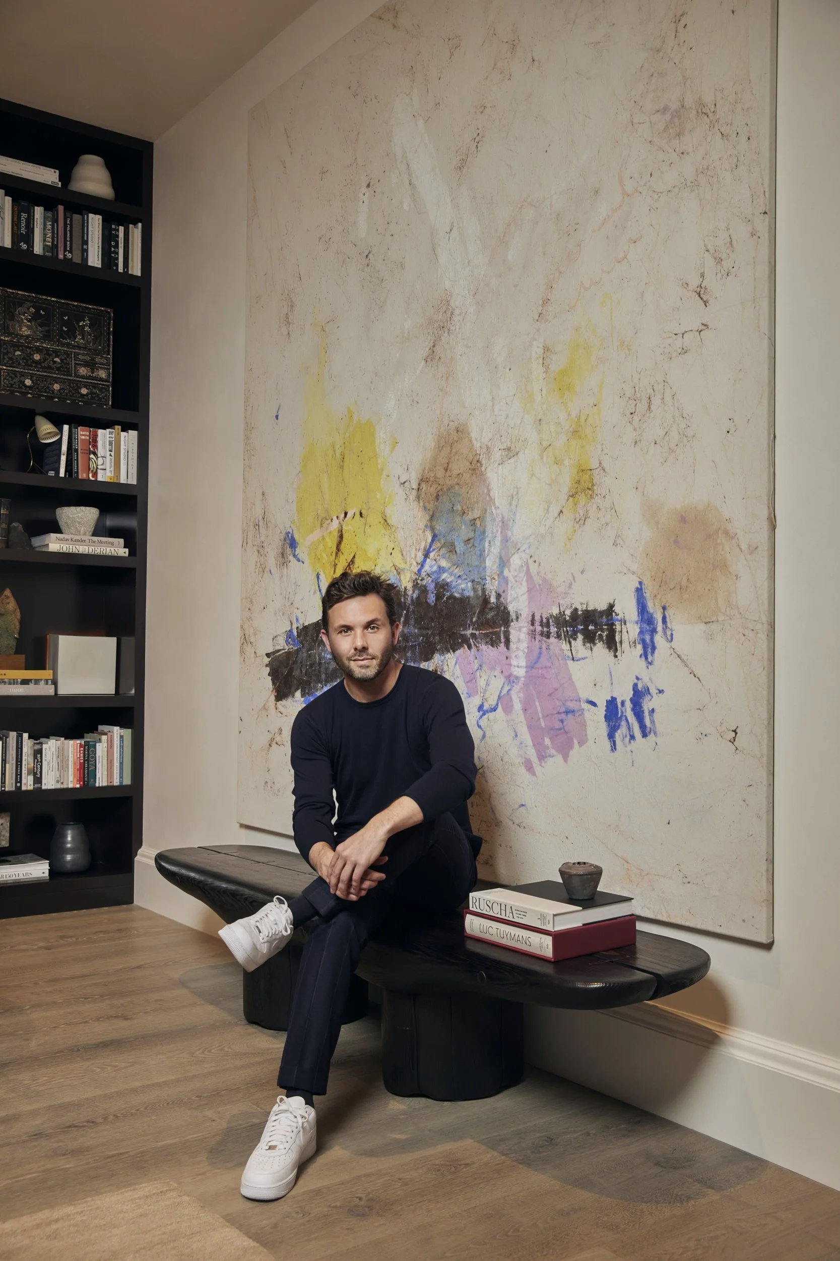 Portrait of Brian Ludlow seated on a black bench before a large abstract painting in soft daylight.