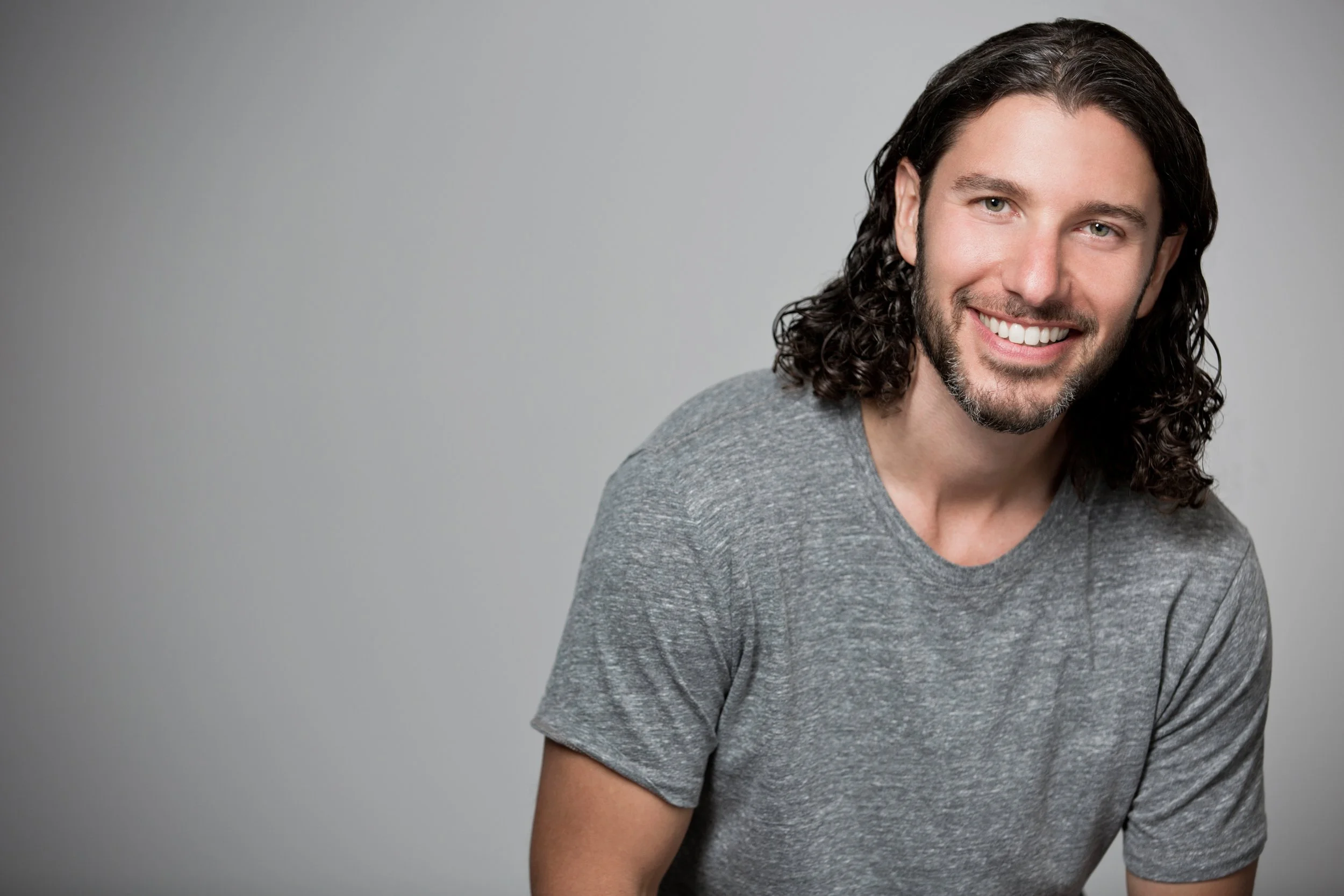 Studio portrait of a smiling man with dark curly hair and a beard against a plain gray backdrop.