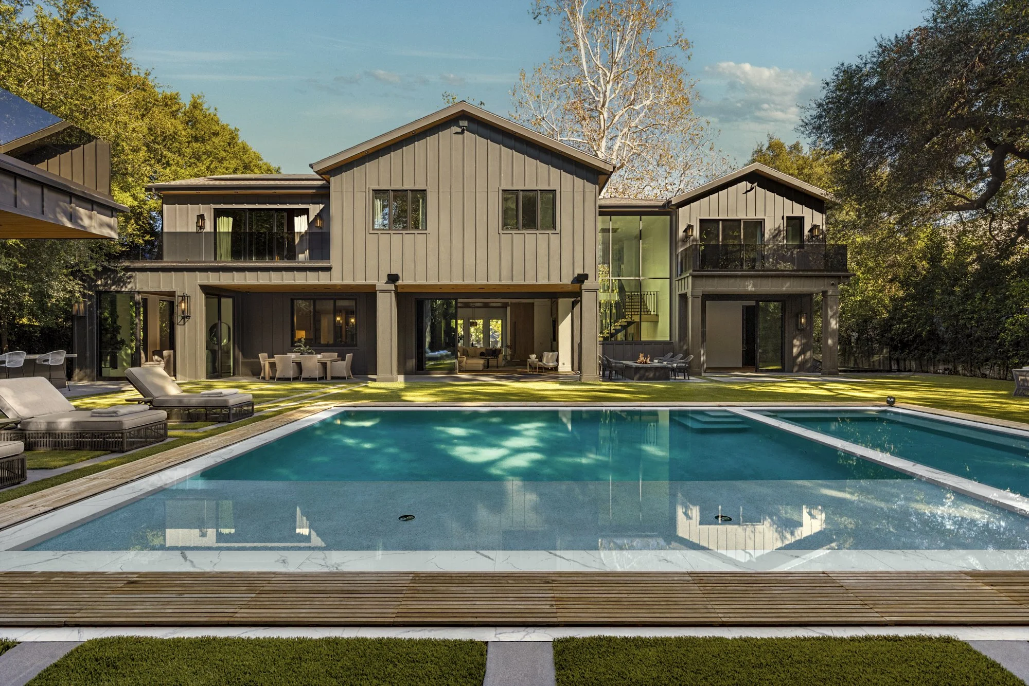 Modern gray residence with a rectangular pool, wood deck, and open glass doors in bright daylight.