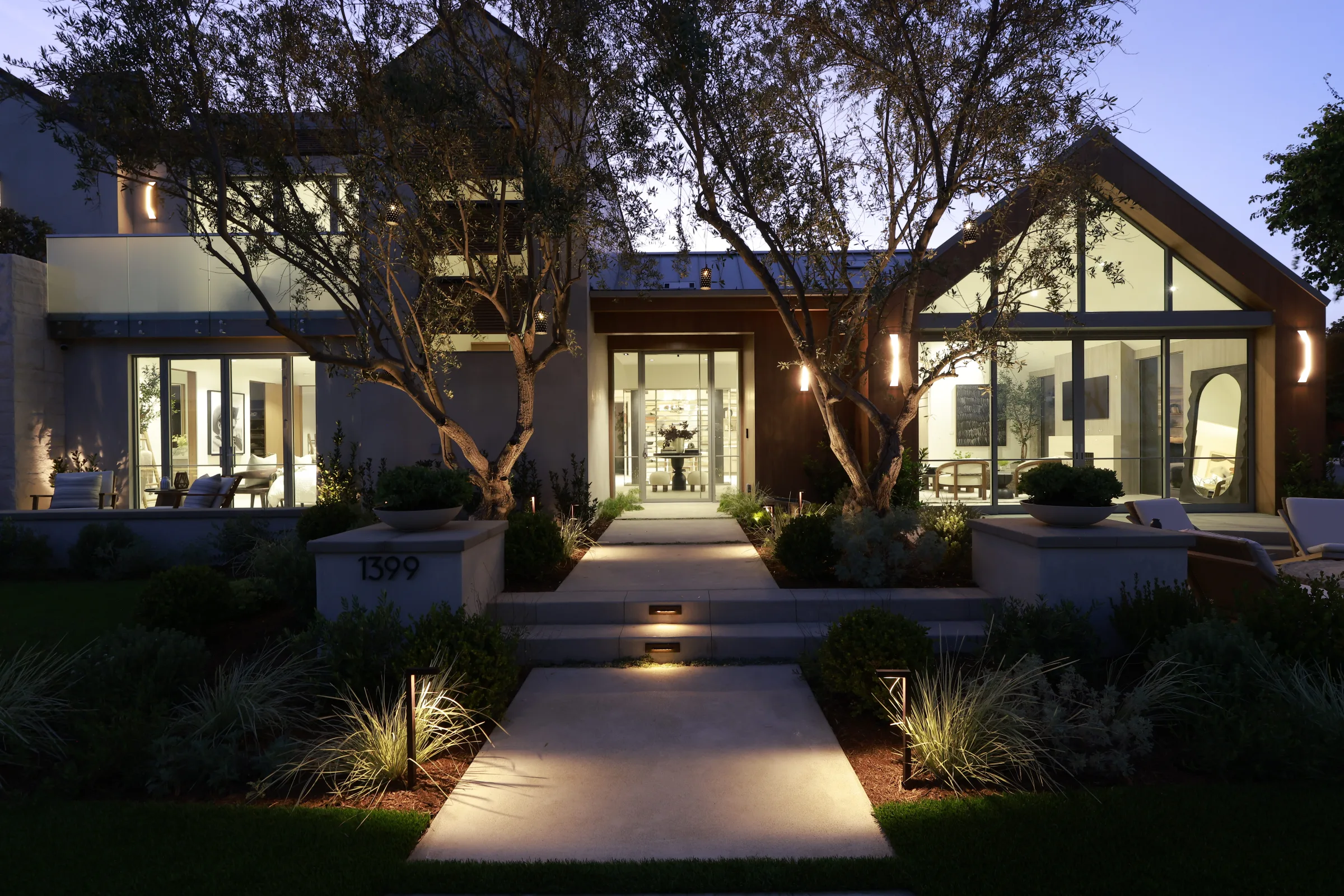 Coastal modern residence at twilight with a lit glass entry, wood gable, and olive trees.