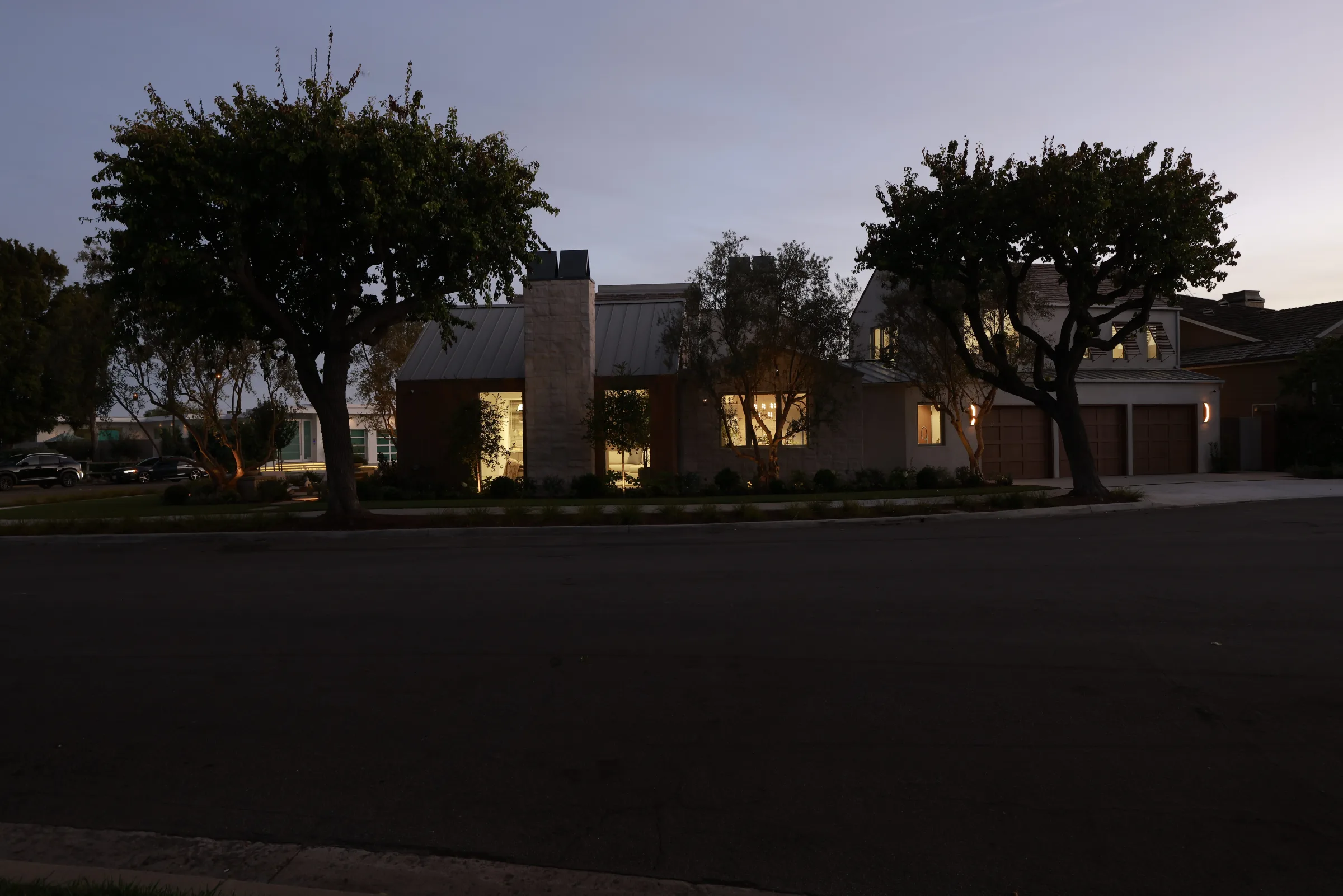 Dusk street facade of a coastal modern house with lit interiors, a stone chimney, and wood garage doors.