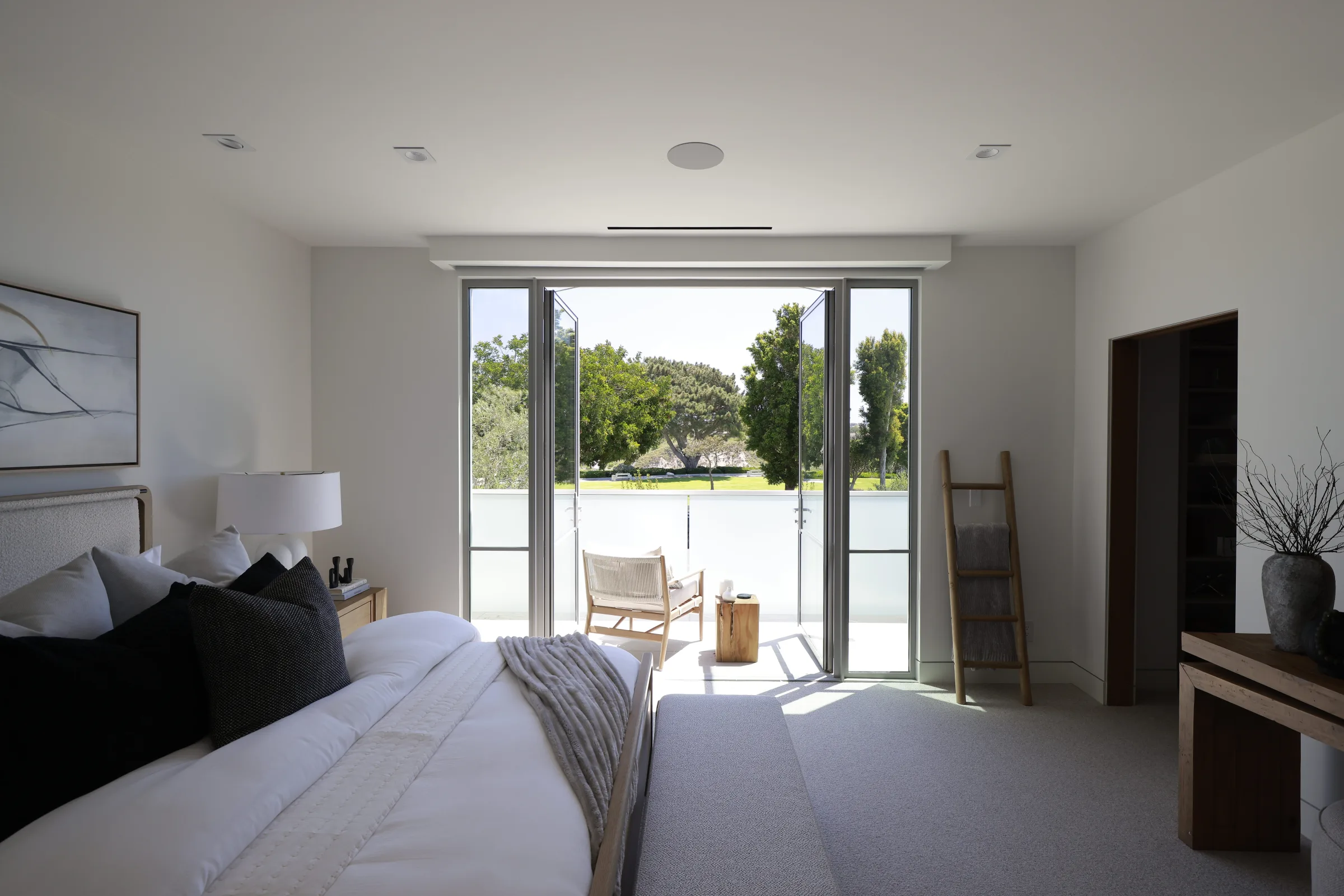 Bedroom with white linens and open balcony doors framed by trees and a clear sky.